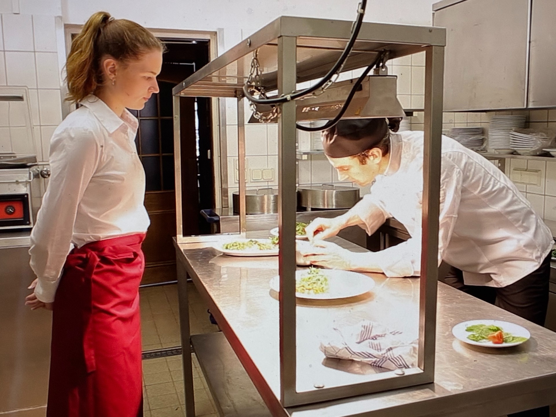 Eine Frau schaut zu, wie ein Mann in der Küche eine Speise garniert. In der Gastronomie braucht man ein Gesundheitszeugnis.